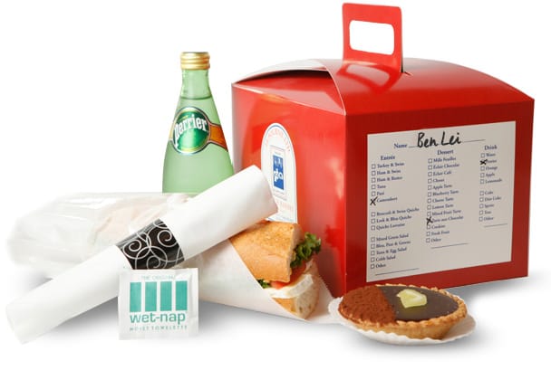 Personalized red lunch box with a menu checklist, wrapped baguette sandwich, Perrier sparkling water, chocolate tart, and rolled napkin with utensils