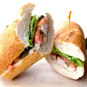 Two baguette sandwich halves filled with lettuce, tomato, and a savory filling on a wooden board