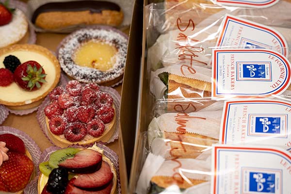 Open catering box of assorted fruit tarts next to individually wrapped baguette sandwiches with Le Fournil labels