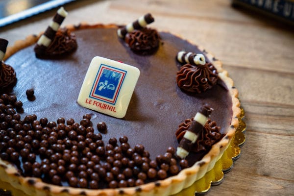 Whole dark chocolate tart topped with crunchy chocolate pearls, piped ganache, and a Le Fournil chocolate plaque